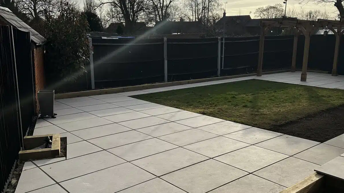 Patio with large grey paving slabs around a small lawn, bordered by a dark fence and pergola