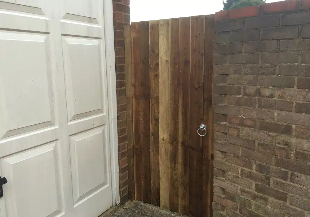 Wooden gate between brick walls beside a white garage door, with a metal ring handle