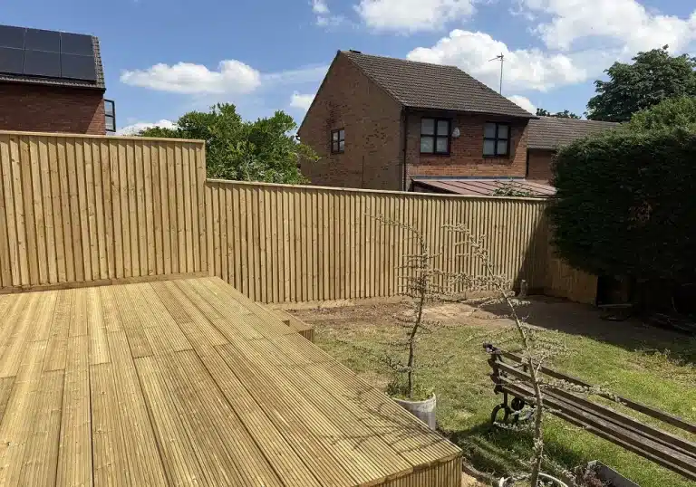 Back garden with wooden deck, fence, lawn and bench under a blue sky